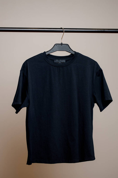 Een leeg zwart T-shirt weegt op een hanger op een neutrale achtergrond. Mock up voor ontwerp.  - Foto, afbeelding
