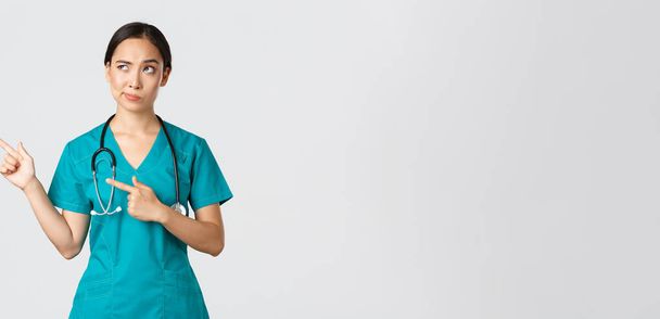 Covid-19, Gesundheitspersonal, Pandemiekonzept. Skeptisch und zweifelhaft hübscher asiatischer Arzt, Krankenschwester im Peeling grinst, zeigt in die linke obere Ecke, blickt mit zögerndem Gesicht, weißer Hintergrund - Foto, Bild