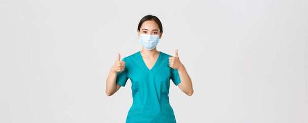 Covid-19 、コロナウイルス病、医療従事者の概念。自信を持って医療マスクやスクラブでプロの女性アジアの医師,承認で親指アップを示します,クリニックでの完璧なサービスを保証 - 写真・画像