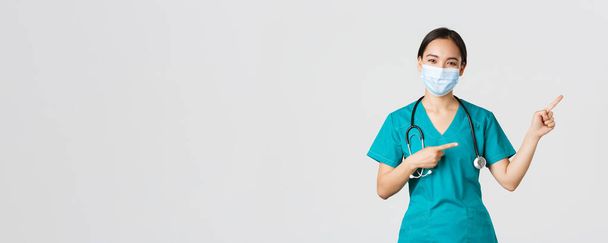 Ковид-19, коронавирусная болезнь, концепция медицинских работников. Приятная азиатка-врач, медицинский работник в халате и маске, указывающий пальцами в правом верхнем углу, показывающий рекламу - Фото, изображение