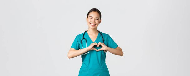 Covid-19, gezondheidswerkers, pandemisch concept. Heerlijk verzorgende aziatische arts, vrouwelijke verpleegster in scrubs tonen hart gebaar en glimlachen, verzorgen van patiënten met liefde, witte achtergrond - Foto, afbeelding