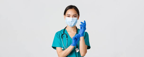 Covid-19, sepelvaltimotauti, terveydenhuollon työntekijöiden käsite. Ammattimainen hymyilevä aasialainen naislääkäri, lääkäri lääketieteellisessä naamiossa ja pensaissa, laittaa kumihanskat ro tutkimus, valkoinen tausta - Valokuva, kuva