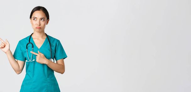 Covid-19, personnel de santé, concept pandémique. Déçu sombre, boudant infirmière asiatique dans les blouses, médecin féminin regardant mécontent, pointant coin supérieur gauche et fronçant les sourcils jugement - Photo, image