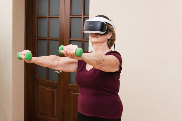 Mujer mayor haciendo ejercicio con pesas de mancuernas en casa usando gafas VR. Anciano activo, entrenamiento de fitness, metaverso.  - Foto, imagen
