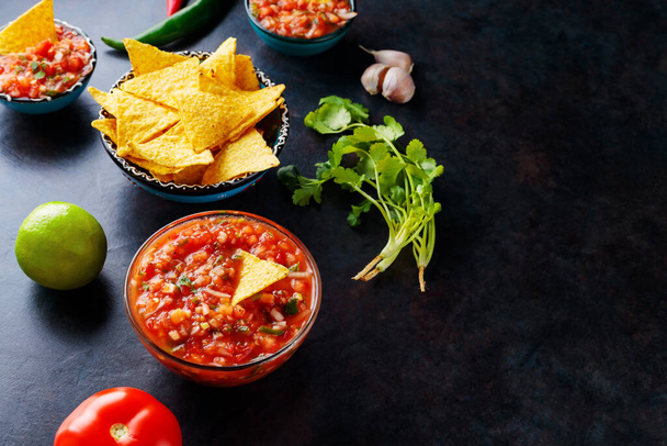 Salsa sauce and ingredients: tomato, onion, garlic, cilantro, lime, pepper. Homemade mexican salsa sauce and nacho chips on a dark background. Top view. Copy space - Fotografie, Obrázek