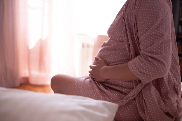 Женщина, страдающая от болей дома, сидящая в постели, трогающая животик и спину, ожидающая родов у женщины - Фото, изображение