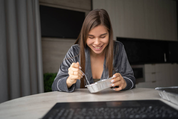 Happy woman enjoying a delicious takeaway lunch at home. A beautiful girl sits at a table and eats from a food container. Food delivery service concept. makes a video call on a laptop. webcam view - Foto, afbeelding