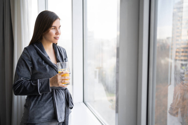people and vacation concept happy caucasian woman drinking citrus juice or smoothie at home standing near window in casual clothes in morning. copy space - Φωτογραφία, εικόνα