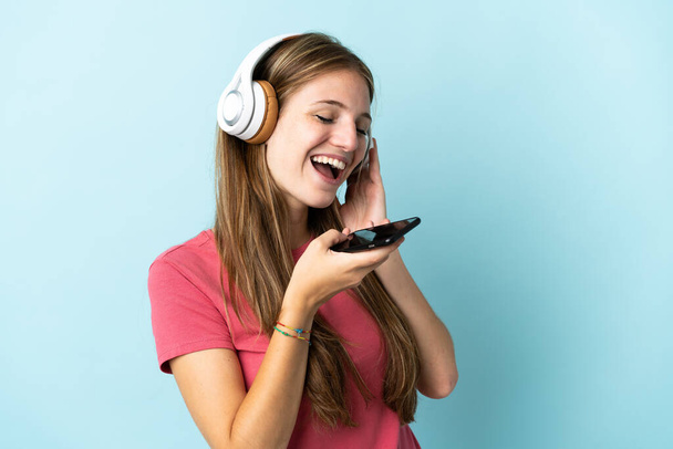 Fiatal kaukázusi nő elszigetelt kék háttér hallgat zenét egy mobil és énekel - Fotó, kép