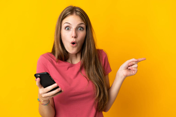 Молодая женщина, использующая мобильный телефон изолирован на желтом фоне удивлен и указатель стороны - Фото, изображение