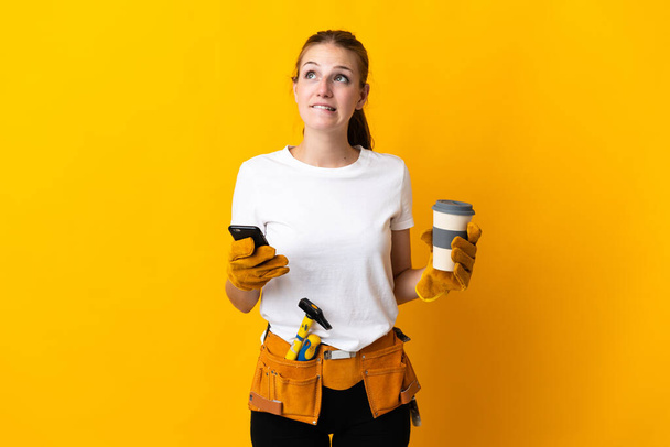 黄色の背景にコーヒーを持って隔離された若い電気技師の女性は何かを考えながら移動する - 写真・画像