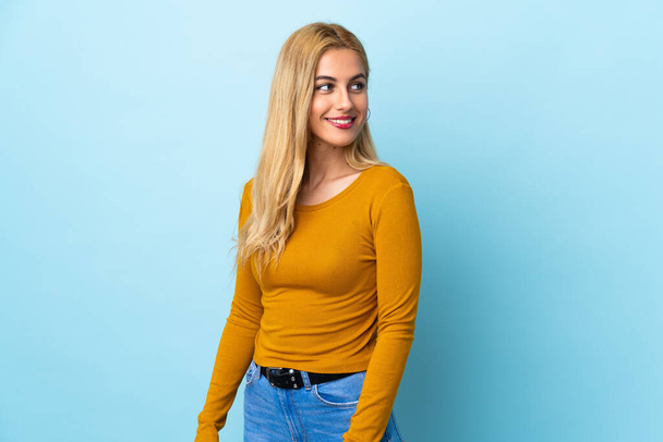 Junge uruguayische blonde Frau vor isoliertem blauen Hintergrund, die zur Seite schaut und lächelt - Foto, Bild