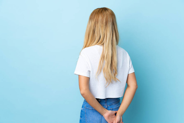 Mladý Uruguayský blondýnka žena přes izolované modré pozadí v zadní poloze a ohlíží - Fotografie, Obrázek
