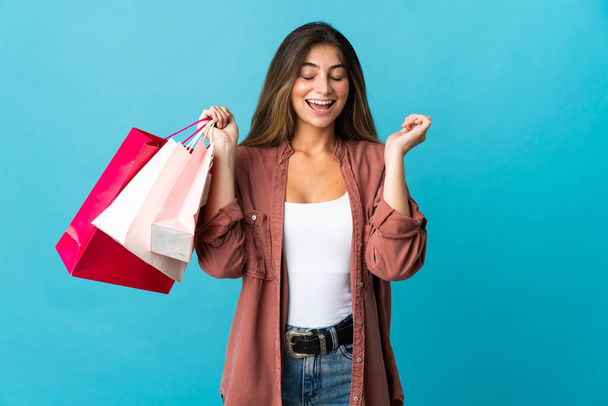 Young caucasian woman isolated on blue background holding shopping bags and smiling - Fotoğraf, Görsel