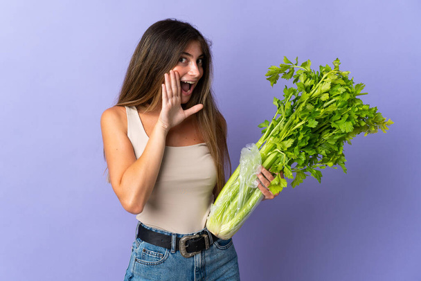 Young woman holding a celery isolated on purple background whispering something - Photo, Image