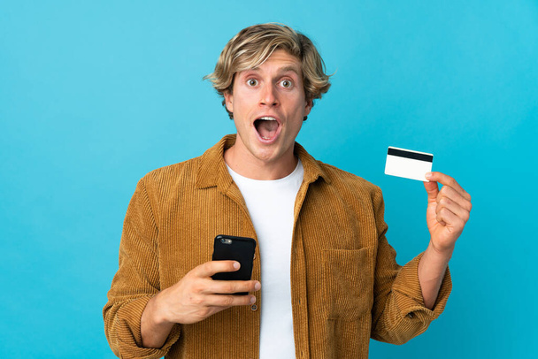 Inglês homem sobre fundo azul isolado compra com o celular e segurando um cartão de crédito com expressão surpresa - Foto, Imagem