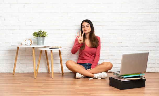 Mujer joven con un ordenador portátil sentado en el suelo en el interior feliz y contando cuatro con los dedos - Foto, imagen