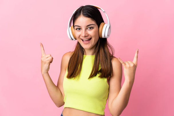 Молодая белая женщина, изолированная на розовом фоне, слушает музыку, делая рок-жест - Фото, изображение