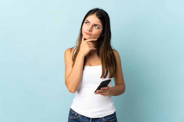 Young caucasian woman using mobile phone isolated on blue background having doubts - Foto, Imagem