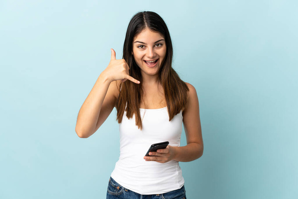 Jonge blanke vrouw met behulp van mobiele telefoon geïsoleerd op blauwe achtergrond het maken van telefoon gebaar. Bel me terug. - Foto, afbeelding