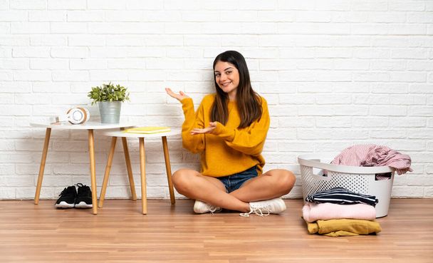 Young woman sitting on the floor at indoors with clothes basket extending hands to the side for inviting to come - Foto, Bild