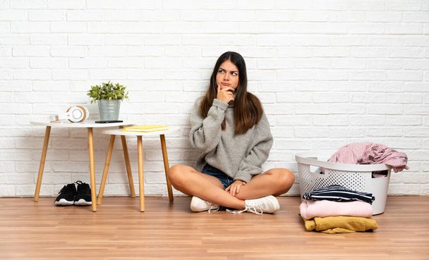 Giovane donna seduta sul pavimento in casa con cesto di vestiti avere dubbi e con espressione faccia confusa - Foto, immagini