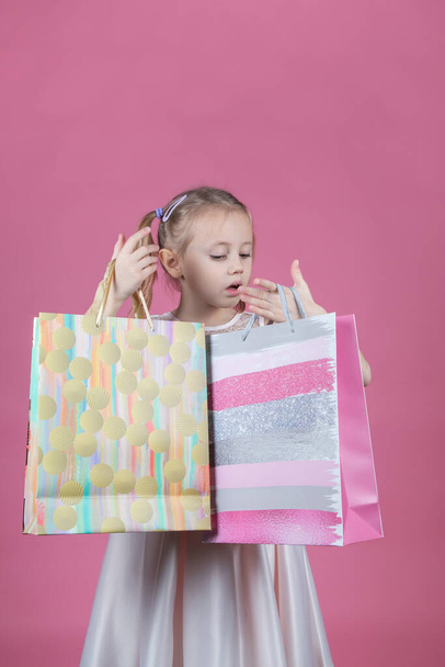 Shopping little caucasian girl in a party dress on pink background holding shopping bags - Foto, imagen