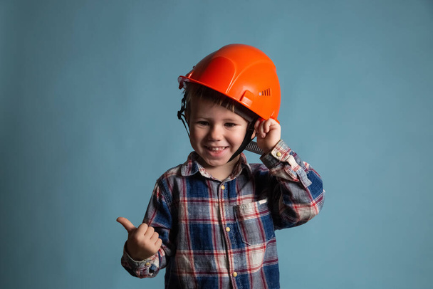 Portrait of cute little boy in a protective orange helmet over blue background showing thumb up. Concept of Construction and repair. - Fotó, kép