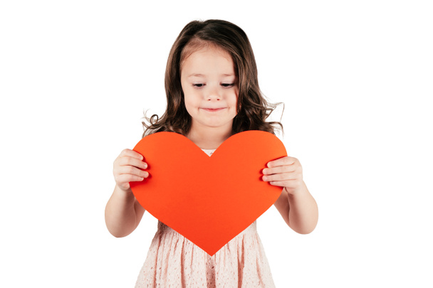 Cute little girl with long curly hair hold in hands big red paper heart lowering eyes forcing smile on white background. - Photo, Image