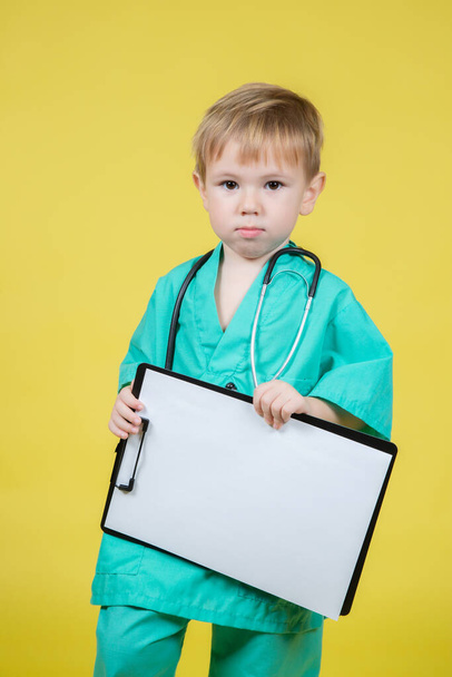 Portrait of little caucasian kid dressed in doctors green coat holds tablet in hand isolated on yellow - Zdjęcie, obraz