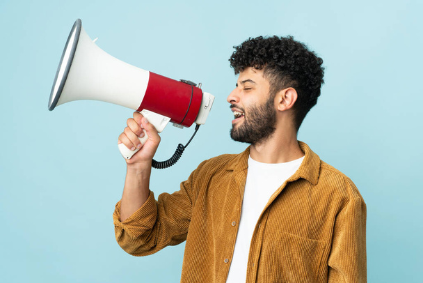 Young Moroccan man isolated on blue background shouting through a megaphone to announce something in lateral position - Photo, Image