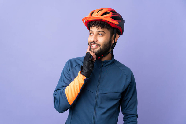 紫の背景に孤立した若いモロッコ人のサイクリストは、上を見上げながらアイデアを考え - 写真・画像