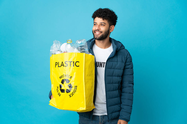 Young Moroccan man holding a bag full of plastic bottles to recycle over isolated background looking side - Фото, изображение