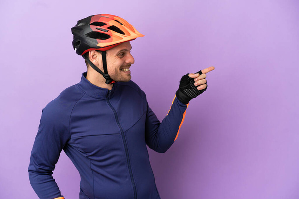 Joven ciclista brasileño aislado sobre fondo púrpura apuntando con el dedo hacia un lado y presentando un producto - Foto, Imagen