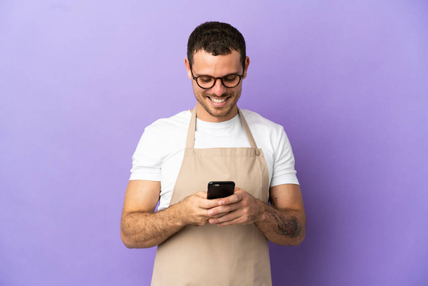 Camarero de restaurante brasileño sobre fondo púrpura aislado enviando un mensaje con el móvil - Foto, Imagen