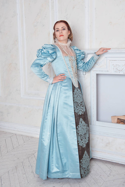 Beautiful young woman in medieval style blue dress stand in the room near fireplace - Foto, Bild