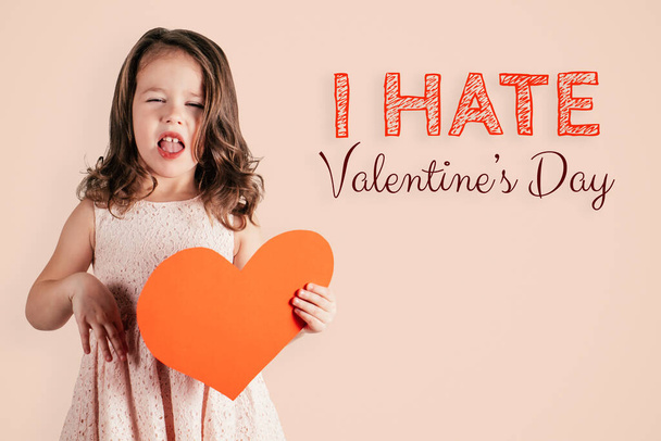 Portrait of little cute girl with long curly hair hold in hand big red paper heart showing disgust on peach background. - Фото, изображение