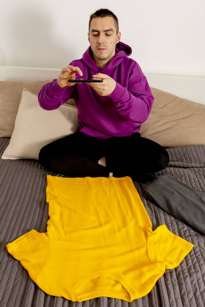 Hombre con capucha violeta usando tableta digital, toma una foto de su ropa vieja para venderlos en línea. Vender en el sitio web, e-commerce. Reutilización, concepto de segunda mano. Consumidor consciente, estilo de vida sostenible - Foto, imagen