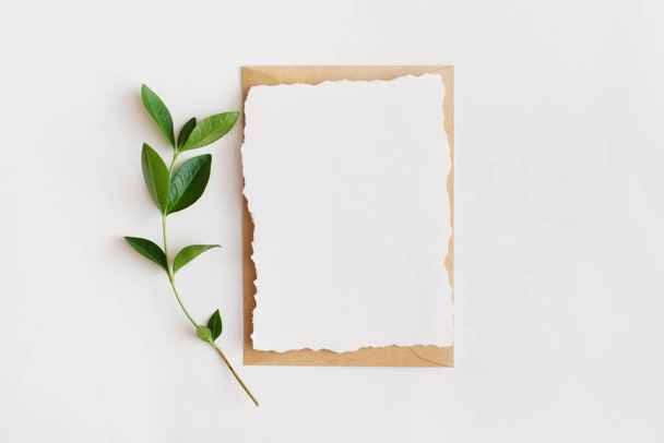 Mock-up of blank paper card with green leaves and envelope on white background. Flat lay, top view with copy space for text - Photo, Image