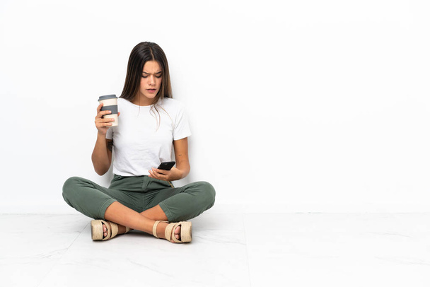 Adolescente chica sentada en el suelo sosteniendo café para llevar y un móvil - Foto, imagen