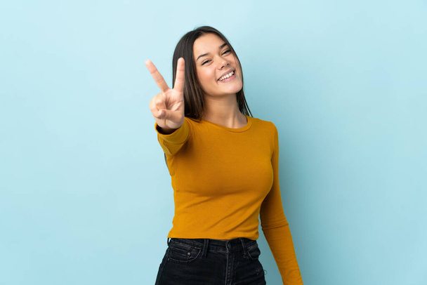 Teenager girl isolated on blue background smiling and showing victory sign - Photo, image