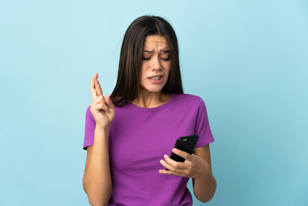 Teenager girl isolated on pink background using mobile phone with fingers crossing - Fotoğraf, Görsel