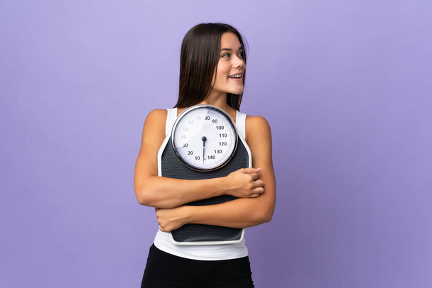 Teenager girl isolated on purple background with weighing machine and looking side - Fotoğraf, Görsel