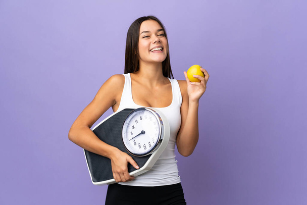 Teenager girl isolated on purple background with weighing machine and with an apple - Photo, Image