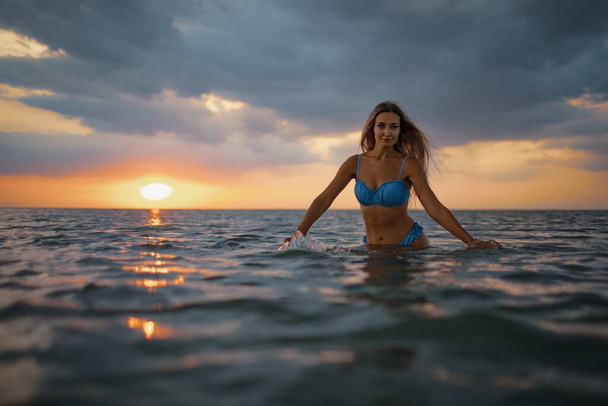 A girl with blond hair in a blue swimsuit splashes to the sides while sitting in an estuary on a sunset background - Foto, Bild