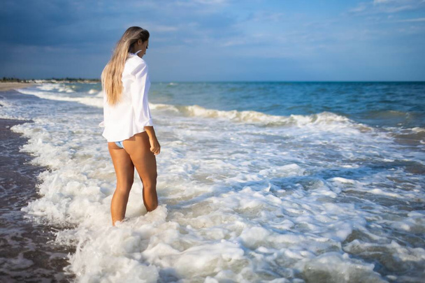 A slender girl in a gentle blue swimsuit and shirt walks along the sandy beach near the blue sea - Foto, afbeelding