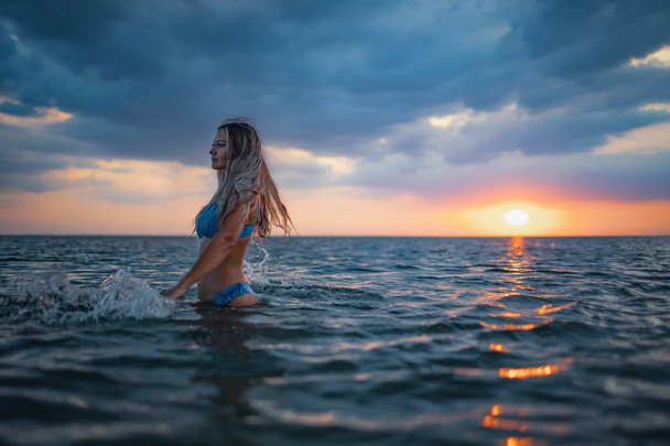 A girl with blond hair in a blue swimsuit splashes to the sides while sitting in an estuary on a sunset background - Фото, зображення