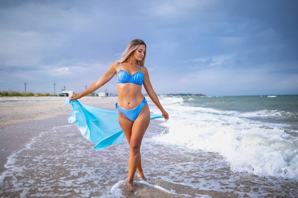 Dziewczyna w blond włosach w niebieskim stroju kąpielowym i jasnym szale spaceruje po plaży - Zdjęcie, obraz