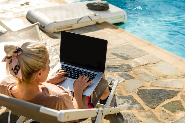 Nous travaillons même en vacances, attrayant jeune femme se trouve sur une chaise longue près de la piscine avec un ordinateur portable, femme utilise un ordinateur portable en vacances pour le travail, travailler en ligne - Photo, image