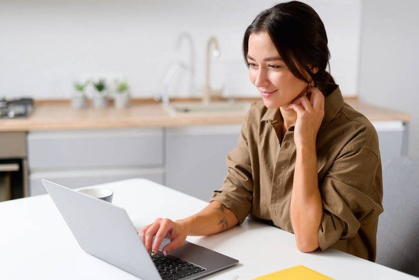 Cheerful asian girl using laptop for remote work or home leisure while sitting in the table at home. Side view of the nice girl looking at the screen - Fotografie, Obrázek